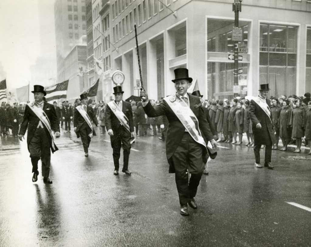 Grand Marshal Malcolm Wilson walks in St. Patrick's Day Parade down a New York City street, with other men marching behind him wearing sashes.