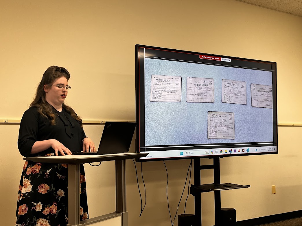 White woman with 1940s hairstyle presenting at a podium with a screen next to her showing World War II ration books