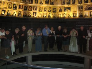 People gather in a semicircle under a display of photographs of Holocaust victims