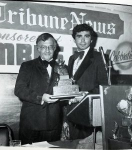 Image of a young man presenting a trophy to an older man, in front of a banner reading "Tribune News"