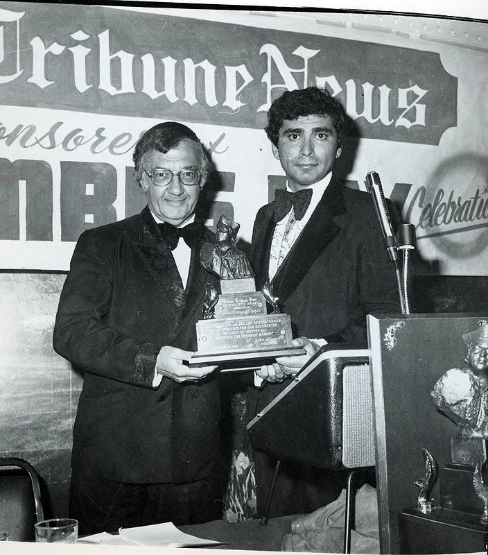 Image of a young man presenting a trophy to an older man, in front of a banner reading "Tribune News"