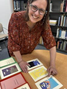 Woman in red shirt standing over a table of illustrated pages of the book A Humument by Tom Phillips.
