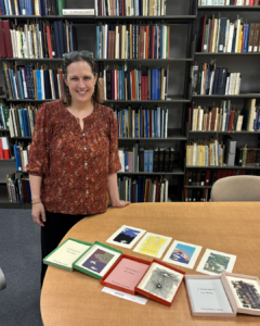 Dr. Sarah Ponichtera stands next to a table with illustrated pages of a book. in the backround there are bookshelves filled with books.