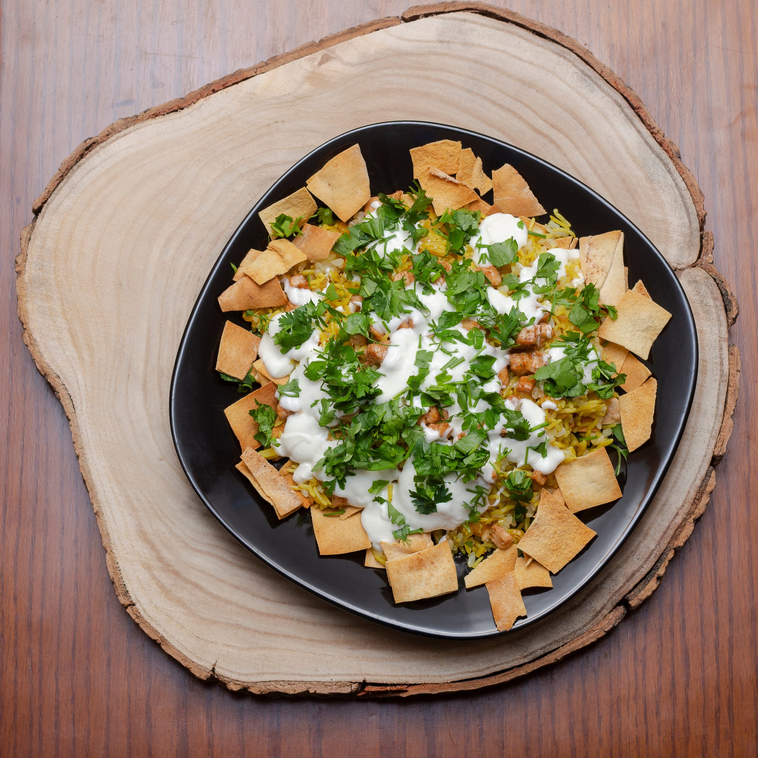 Fatta, rice, chicken, yogurt, roasted bread pieces and parsley, in black plate, on wooden log