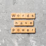 The image features wooden letter tiles arranged on a textured gray concrete background, spelling out "WORDS HAVE POWER," with the contrast between the rough surface and the structured tiles emphasizing the significance of language and its influence.