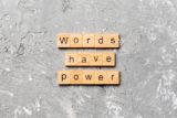 The image features wooden letter tiles arranged on a textured gray concrete background, spelling out "WORDS HAVE POWER," with the contrast between the rough surface and the structured tiles emphasizing the significance of language and its influence.
